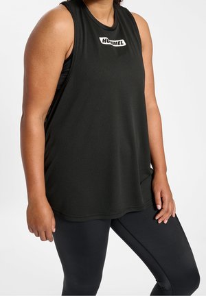 Osoba ubrana w czarny sportowy top bez rękawów z logo "Hummel" oraz czarne legginsy, stojąca na tle jednolitej białej ściany.