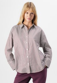 Blonde vrouw in een losse bruine en witte verticaal gestreepte blouse met knopen, handen in de zakken, gecombineerd met donkerpaarse broek, witte achtergrond.