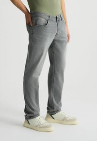 Jeans de mezclilla gris claro con corte recto, cinco bolsillos y cierre de botón, combinados con zapatillas blancas y verdes. Textura suave.
