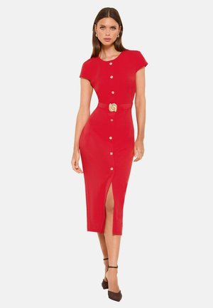 Frau trägt ein eng anliegendes rotes Kleid mit kurzen Ärmeln, goldenen Knöpfen vorne, einem Gürtel mit goldener Schnalle und einem Schlitz vorne, kombiniert mit schwarzen High Heels.