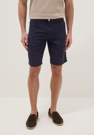 Mann, der marineblaue Shorts und dunkelbraune Slipper trägt, steht vor einem schlichten hellen Hintergrund.