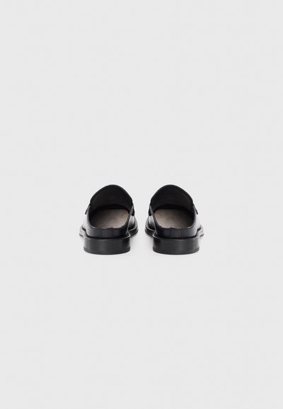 Sapatos slip-on pretos em couro com uma textura suave, bico redondo, salto baixo e design minimalista, destacando os detalhes do forro interior.