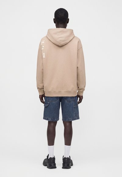 Homme de dos portant un sweat à capuche beige avec un texte blanc sur la manche, un short en jean bleu, des chaussettes blanches et des baskets noires.
