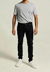 Homme portant un t-shirt à manches courtes gris clair, un pantalon slim noir et des baskets blanches, debout sur un fond uni.