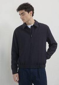 Giacca zip-up blu navy con colletto, cintura elastica e maniche lunghe. Realizzata in tessuto liscio con un design aderente.