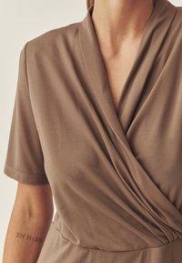 Braun Wickelkleid mit kurzen Ärmeln, aus weichem, strukturiertem Stoff. Verfügt über ein plissiertes Design an der Brust und eine taillierte Taille, die die Form betont.