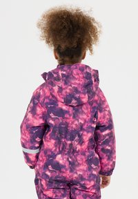 Barn iført en pink og lilla galakse-mønstrede vinterdragt med hætte, set fra ryggen, stående mod en ensfarvet hvid baggrund.