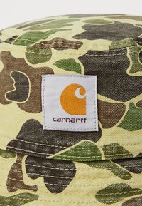 Camo bucket hat in yellow, green, and brown fabric. Features a white tag with an orange logo stitched on a textured surface.