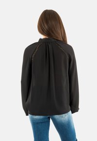 Femme aux longs cheveux bruns portant un blouse noire avec des détails en dentelle sur les épaules et un jean bleu, se tenant de dos à la caméra.