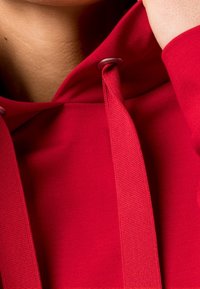 Czerwona bluza z kapturem o miękkiej teksturze, z regulowanym kapturem i metalowymi oczkami. Materiał jest gładki z lekkim rozciągnięciem.