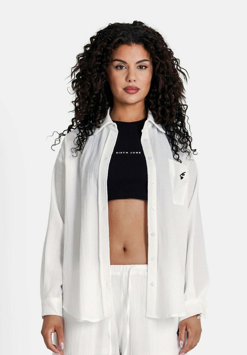Hvid, tekstureret skjorte med afslappet pasform og lange ærmer, med en frontlomme. Båret over en sort cropped top.