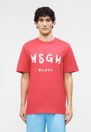 Νεαρός με σγουρά μαύρα μαλλιά φοράει κόκκινο t-shirt MSGM Milano και ανοιχτόχρωμο μπλε παντελόνι, στέκεται μπροστά σε απλό ανοιχτό γκρι φόντο.