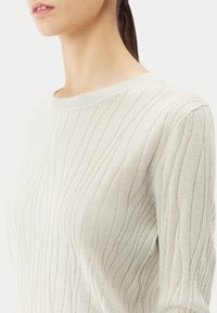 Pull en tricot beige clair avec une texture côtelée, col rond et léger éclat, mettant en valeur des lignes verticales et une silhouette ajustée.