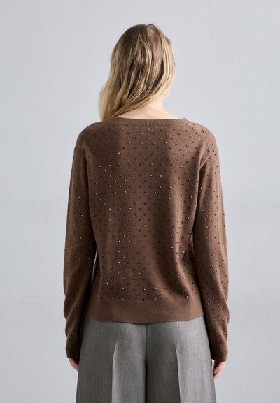 Kobieta z długimi blond włosami nosi brązowy sweter z małymi srebrnymi ćwiekami i szerokie szare spodnie, stojąc na jednolitym tle.