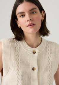Gilet en tricot crème avec un col rond, présentant un motif de tricot en relief et deux boutons foncés à l'avant. Tissu texturé, design sans manches.