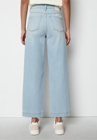 Osoba v světle modrých širokých džínách a bílých teniskách, stojící s výhledem k bílému pozadí.