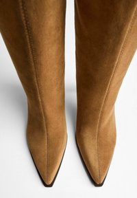 Bottes en suédine beige montantes avec un bout pointu et une texture lisse, présentant des coutures verticales sur les côtés et une semelle foncée contrastante.