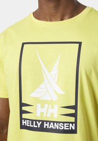 Camiseta de algodón amarilla con un logo blanco y un diseño de caja negra, con gráficos angulares y el texto "HELLY HANSEN" en la parte inferior.