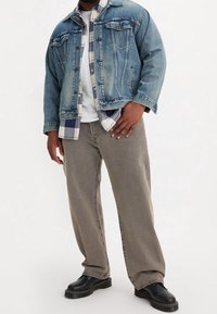 Kurtka dżinsowa z wyblakłym niebieskim wykończeniem i zapięciem na guziki, noszona na wierzchu koszuli w kratę flanelową, zestawiona z beżowymi spodniami o luźnym kroju i czarnymi butami.