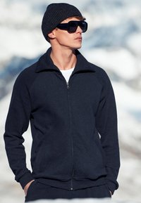 Dunkelblaues Zip-Up Sweatshirt mit hohem Kragen, gerippten Bündchen und passender Hose, getragen mit einer Strickmütze und übergroßen Sonnenbrillen.