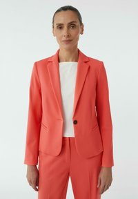 Koraalgekleurde blazer met notch-lapels, enkele knoopsluiting en twee voorkpresas, gecombineerd met een witte top en bijpassende broek.