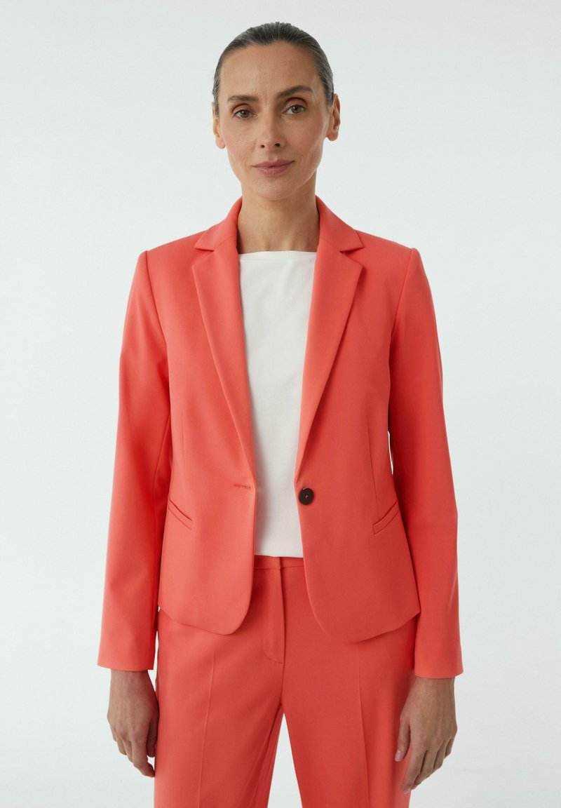Koraalgekleurde blazer met notch-lapels, enkele knoopsluiting en twee voorkpresas, gecombineerd met een witte top en bijpassende broek.