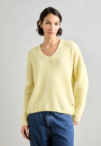 Gelber, V-Ausschnitt, übergroßer Pullover aus weichem Stoff mit geripptem Muster. Kombiniert mit blauen, lässig geschnittenen Jeans. Subtle Akzentdetail am Saum.