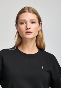 Svart sweatshirt i mjukt material, med rund halsringning och långa ärmar. En liten vit logotyp är broderad på vänster bröstområde.
