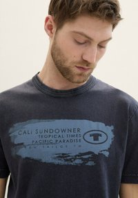 Mørkeblå bomulds t-shirt med grafisk tryk i lyseblå med teksten: "CALI SUNDOWNER TROPICAL TIMES PACIFIC PARADISE." Korte ærmer.