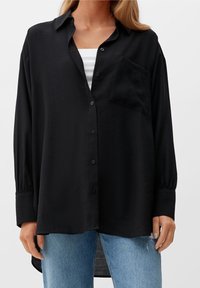 Camicia nera a maniche lunghe con bottoni, realizzata in un tessuto morbido, dotata di una tasca sul petto e dal taglio morbido, abbinata a jeans azzurri chiari.