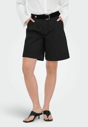 Žena v čiernych ľahko prispôsobených šortkách s opaskom, bielej košeli s dlhým rukávom a čiernych sandáloch na podpätkoch, stojaca s rukami v vreckách.