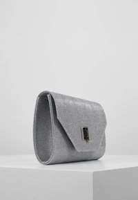 Sølv metallisk clutch-taske med en struktureret overflade, kuvertdesign, magnetisk lukning og en lille rektangulær hardwaredetalje på fronten.