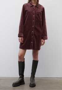 Burgundy corduroy-mekko pitkillä hihoilla, nappikiinnitys edessä ja rintatasku. Yhdistetty polveen ulottuviin mustiin kumisaappaisiin.
