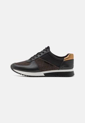 Niedriger schwarzer Sneaker aus strukturiertem Stoff, mit Lederakzenten, weißer Zwischensohle und metallisch goldenem Fersen-Detail, seitlich auf weißem Hintergrund dargestellt.