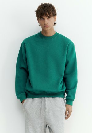 Junge Person mit kurzen braunen Haaren, die einen grünen Pullover und hellgraue Jogginghosen trägt, steht mit den Händen in den Taschen.