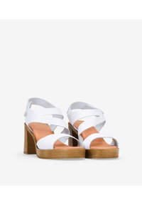 Sandalias de tacón blanco hechas de un material texturizado, con diseño de tiras cruzadas, punta abierta y un taco de madera color marrón claro.