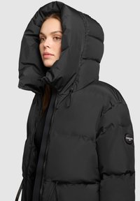 Crna puffer jakna s velikim kapuljačom, vidljivim zatvaračem i elastičnim vezicama. Tekstuirani, kviltani dizajn s logom na rukavu.