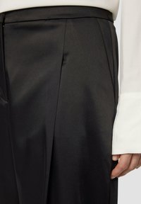 Schwarze Satin-Hosen mit glatter Textur, ausgestattet mit vorderen Falten und einem Reißverschluss. Hand sichtbar, trägt ein langärmeliges weißes Hemd.