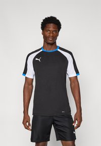 Cămășă sport neagră cu accente albastre și albe, având o textură din plasă pe mâneci și logo pe piept. Asortată cu pantaloni scurți negri.