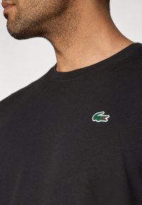 Czarna bawełniana koszulka z zaokrąglonym dekoltem, z małym zielonym logo aligatora na lewej stronie klatki piersiowej. Gładka faktura.