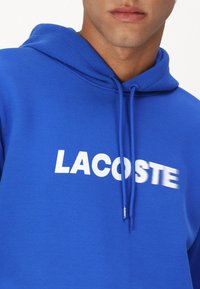 Hoodie bleu en tissu texturé avec un logo "LACOSTE" blanc sur le devant et des cordons pour la capuche.