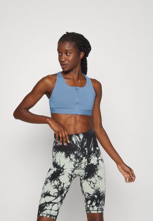 Top cropped atlético azul claro com um fecho na frente e alças largas, combinado com calções de cintura alta em tie-dye verde e preto.