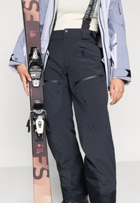 Zwarte skibroek met verstelbare banden, ritszakken en textuursecties, gecombineerd met een ski en bindingen. Het materiaal lijkt duurzaam.