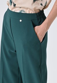 Groene, wide-leg broeken met een gladde textuur, elastische tailleband en zijzak. Een klein zilveren knoopdetail is zichtbaar nabij de tailleband.