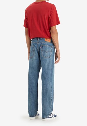 Persoon draagt een rood t-shirt, blauwe rechte spijkerbroek met hoge taille en achterzakken, en witte sneakers met blauwe accenten, staand met de rug naar voren.