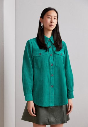 Camicia in corduroy verde con chiusura a bottoni, due tasche sul petto e maniche lunghe. Presenta bottoni neri e una texture liscia.