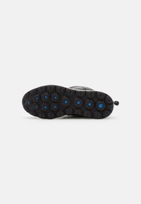 Suela de bota de goma negra con patrones de tracción circulares e insertos azules, vista desde abajo sobre un fondo blanco.
