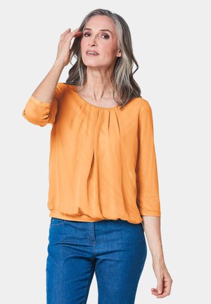 Orange Bluse mit Dreiviertelärmeln, rundem Ausschnitt und plissierter Vorderseite. Kombiniert mit blauen Jeans in einem lässigen Schnitt.