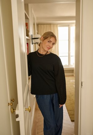 Jeune femme aux cheveux blonds attachés porte un haut noir à manches longues et un jean bleu, debout dans un cadre de porte à l'intérieur d'une pièce lumineuse.