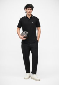 Tricou polo negru cu accente albe, pantaloni negri largi și adidași în tonuri neutre. Modelul ține în mână un mic pouch gri pentru accesorii.